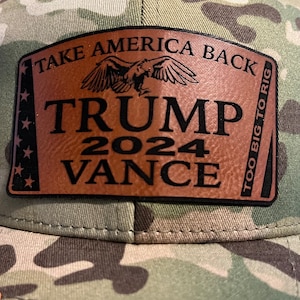 Può includere: Un cappello da camionista mimetico con una toppa in pelle marrone che dice "Take America Back" con un'aquila sopra il testo, "Trump 2024 Vance" sotto l'aquila e "Too Big To Rig" sul lato destro della toppa.