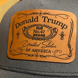 Puede incluir: Un parche de cuero marrón con texto negro que dice "Donald Trump Old No. 47 Brand United States of America". El parche está unido a una gorra de camionero gris.