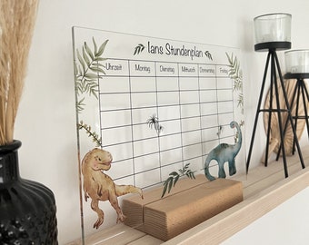 Peut inclure: Un tableau d'horaire hebdomadaire en acrylique transparent sur le thème des dinosaures. Le tableau est composé d'une grille avec des colonnes pour chaque jour de la semaine et des lignes pour les créneaux horaires. Le tableau est décoré de feuilles vertes et de deux dinosaures, un brun et un bleu.