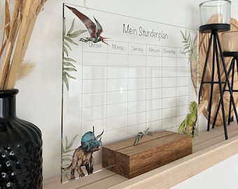 May include: A clear acrylic weekly planner with dinosaur illustrations and the text "Mein Stundenplan". The planner features a grid for each day of the week, with a wooden block base. Decorative plants and a black vase are also visible.