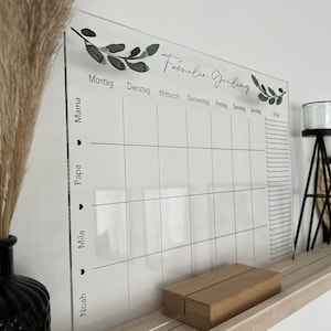 Puede incluir: Un calendario familiar de acr&iacute;lico transparente con el texto "Familie Gerling" y columnas diarias. El calendario tiene una base de madera y est&aacute; decorado con detalles de hojas verdes. Los nombres "Mama", "Papa", "Mila" y "Noah" est&aacute;n listados a la izquierda.