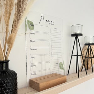 Könnte beinhalten: Ein transparenter, wöchentlicher Menüplaner aus Acryl mit einem Holzständer. Der Planer hat ein schlichtes Design mit einer schwarzen Überschrift "Menu der Woche" und einer Liste der Wochentage: Montag, Dienstag, Mittwoch, Donnerstag, Freitag, Samstag und Sonntag. Jeder Tag hat Platz zum Eintragen von Mahlzeiten und einen Bereich für Notizen.