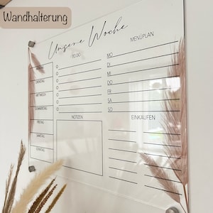 Puede incluir: Un planificador semanal de acr&iacute;lico transparente con la inscripci&oacute;n "Unsere Woche". El planificador tiene secciones para listas de tareas, un men&uacute;, notas y listas de compras. Est&aacute; decorado con acentos florales secos y montado con herrajes plateados.