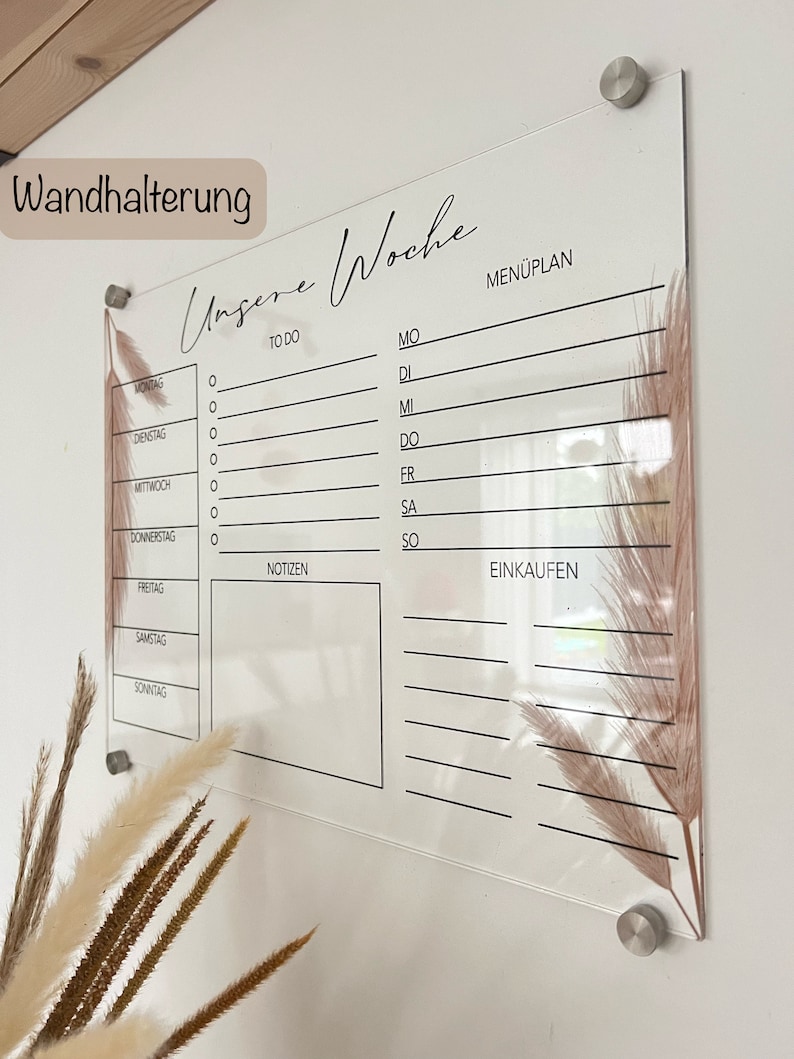 Puede incluir: Un planificador semanal de acr&iacute;lico transparente con la inscripci&oacute;n "Unsere Woche". El planificador tiene secciones para listas de tareas, un men&uacute;, notas y listas de compras. Est&aacute; decorado con acentos florales secos y montado con herrajes plateados.