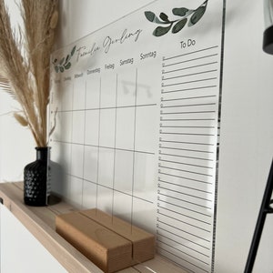 Puede incluir: Un calendario de acr&iacute;lico transparente con una base de madera. El calendario presenta un dise&ntilde;o semanal con los d&iacute;as de la semana y una secci&oacute;n "To Do". Acentos decorativos de hojas verdes y el texto "Familie Gerling" est&aacute;n impresos en la parte superior.