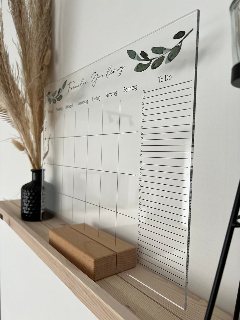Puede incluir: Un calendario de acr&iacute;lico transparente con una base de madera. El calendario presenta un dise&ntilde;o semanal con los d&iacute;as de la semana y una secci&oacute;n "To Do". Acentos decorativos de hojas verdes y el texto "Familie Gerling" est&aacute;n impresos en la parte superior.