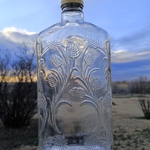 May include: Clear glass bottle with embossed floral design and a gold-colored cap. The bottle has a rectangular shape with rounded shoulders and a detailed pattern of stylized flowers. The background shows a cloudy sky and a wooden fence.