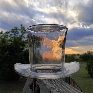 May include: A clear glass top hat-shaped container with a wide brim, resting on a wooden fence. The sky in the background displays a colorful sunset with shades of orange, yellow, and blue, creating a striking contrast with the clear glass.