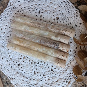 May include: Five glass vials filled with dried botanicals, each sealed with a cork stopper. The vials are arranged on a white, crocheted doily. The contents of the vials vary in color, ranging from light beige to brown, and include delicate, textured elements.