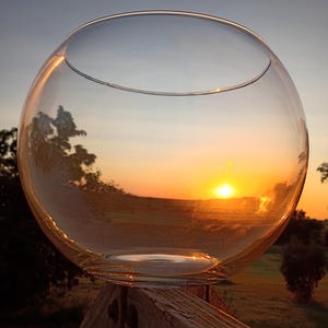 May include: A clear, spherical glass bowl sits atop a wooden structure, reflecting the warm hues of a sunset. The bowl's smooth surface captures the golden light, creating a serene and natural scene.