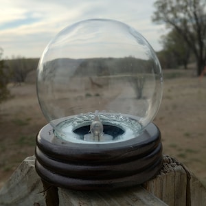 May include: A clear glass globe sits atop a dark brown, tiered wooden base. Inside the globe is a small light bulb. The object is placed on a weathered wooden fence post, with a blurred outdoor background.