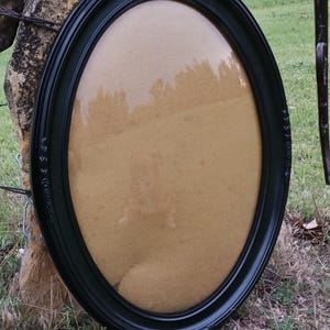 May include: An oval picture frame with a smooth black finish and subtle decorative embossed detailing along the outer edge. The frame features a clear glass front covering a plain tan backing, creating a reflective surface within the oval border.