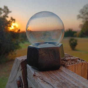 May include: A clear glass orb candle holder with a dark, geometric base, resting on a weathered wooden surface. The background features a sunset with warm orange and yellow hues, creating a serene and natural ambiance.