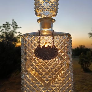 May include: A clear, diamond-cut glass decanter with a stopper, labeled "GIN" on a copper-colored tag. The decanter is illuminated from within, creating a warm glow against a sunset backdrop. The bottle is a decorative item.