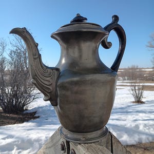 May include: An antique silver-colored teapot with a curved spout and a black handle. The teapot has a decorative design on the spout and a lid with a small knob. The teapot is sitting on a wooden surface outdoors.