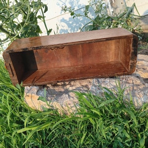 May include: A rectangular, open-top wooden box with a dark brown finish. The box has a simple, rustic design with rounded corners and small handles on the sides. The box is resting on a stone surface, surrounded by green grass and foliage.