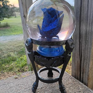 May include: A clear glass orb encases a blue rose, set on a black metal stand. The rose is a deep blue with glitter, and the stand has ornate details. The stand is supported by three curved legs, creating a decorative display.