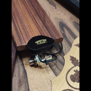 May include: Close-up of a black guitar pickup with a gold-colored jack and a black wire. The pickup has the word "TAP" printed on it in gold letters.