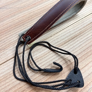 May include: A brown leather guitar strap with a black cord and a hook. The strap is designed to be worn around the neck and has a hook to attach to the guitar.