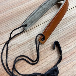 May include: A brown leather camera strap with a gray suede lining and a black adjustable strap. The strap has a black triangular buckle.