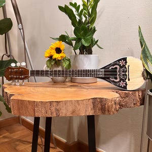 May include: A wooden table with a natural wood top and black metal legs. A bouzouki, a Greek stringed instrument, is resting on the table. A sunflower is in a white vase next to the instrument.
