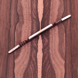 May include: A white and brown guitar bridge saddle with a black inlay. The saddle is on a dark brown wood surface.