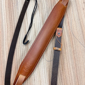 以下が含まれることがあります： 茶色の革製のギターストラップで、黒い革製のストラップと金属製のバックルが付いています。ストラップには、茶色の革製のループと金属製のフックが付いています。