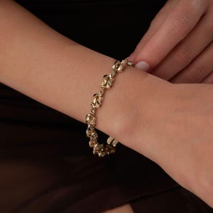 Puede incluir: Un brazalete dorado con un diseño geométrico. El brazalete está hecho de pequeñas formas interconectadas que crean una superficie texturizada. El brazalete se está abrochando en una muñeca.