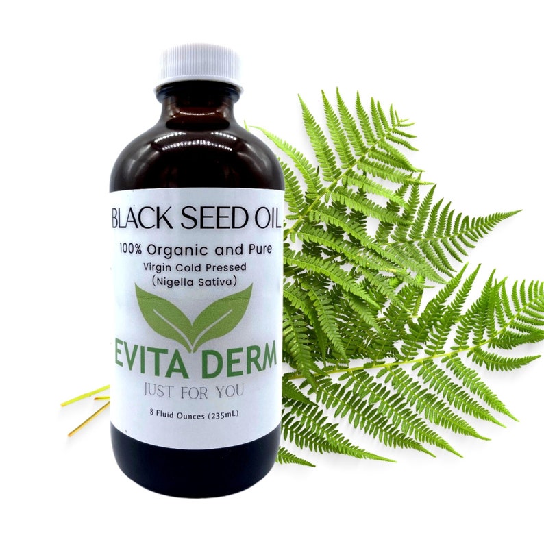 May include: A brown glass bottle with a white label that reads "BLACK SEED OIL, 100% Organic and Pure Virgin Cold Pressed (Nigella Sativa) EVITA DERM JUST FOR YOU 8 Fluid Ounces (235mL)". The bottle is sitting on a white background with green fern leaves behind it.