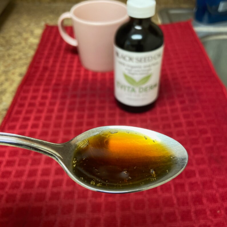 May include: A close-up of a silver spoon filled with a dark amber liquid. The liquid is thick and viscous, and it has a slight sheen. The spoon is resting on a red checkered tablecloth. A bottle of black seed oil is visible in the background.