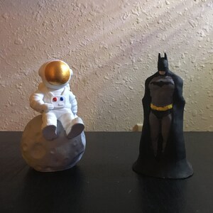 May include: A white astronaut figurine sitting on a gray moon-shaped base, next to a black Batman figurine.