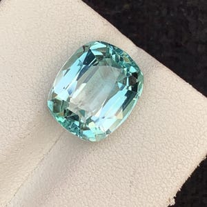 May include: A light blue gemstone with a rectangular cushion cut. The gem is set on a white surface against a dark background. The gemstone has multiple facets that reflect light.