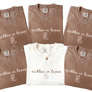May include: A collection of brown and white t-shirts with the phrase "reckless in texas" and a cowboy boot graphic. Each shirt has a silver necklace. The white shirt has a ring graphic.