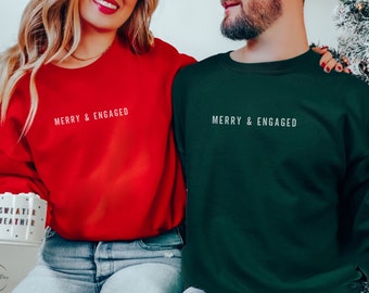 May include: Red and forest green crewneck sweatshirts with the text "MERRY & ENGAGED" in white. The red sweatshirt is paired with light blue jeans. The forest green sweatshirt is paired with light blue jeans. The sweatshirts are perfect for couples.