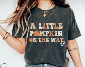 May include: Dark gray t-shirt with the text "A Little Pumpkin On The Way" in white, orange, and tan lettering. The shirt features a pumpkin graphic and baby footprints. The shirt is worn with blue jeans.