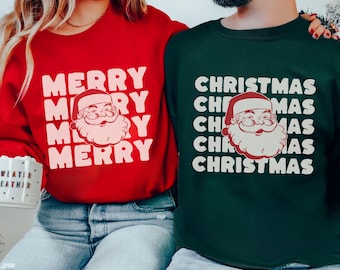 May include: Two sweatshirts, one red and one forest green, with holiday-themed designs. The red sweatshirt says "MERRY" multiple times with a Santa face. The green sweatshirt says "CHRISTMAS" multiple times with a Santa face. Both have white text.