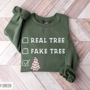 May include: A military green sweatshirt with the text "REAL TREE" and "FAKE TREE" in white. A checkmark is next to a cartoon Christmas tree design. The sweatshirt is on a light wood background.