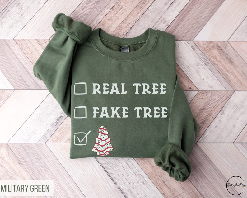 May include: A military green sweatshirt with the text "REAL TREE" and "FAKE TREE" in white. A checkmark is next to a cartoon Christmas tree design. The sweatshirt is on a light wood background.
