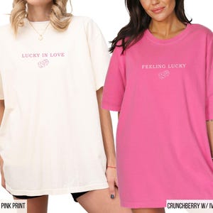 May include: Two oversized t-shirts, one ivory and one pink, with short sleeves. The ivory shirt has the text "LUCKY IN LOVE" and a dice graphic in pink. The pink shirt has the text "FEELING LUCKY" and a dice graphic in ivory.