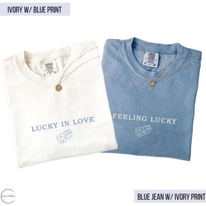 May include: Two t-shirts, one ivory and one blue, with gold necklaces. The ivory shirt says "LUCKY IN LOVE" with dice graphics. The blue shirt says "FEELING LUCKY" with dice graphics. Both shirts are Comfort Colors brand.