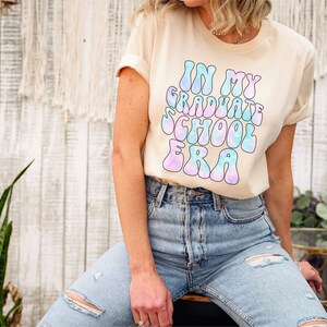 May include: A light beige t-shirt with a tie-dye effect graphic that says "In My Graduate School Era" in a retro font.
