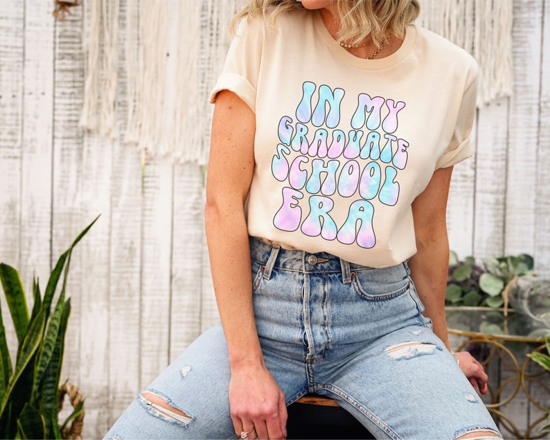 May include: A light beige t-shirt with a tie-dye effect graphic that says "In My Graduate School Era" in a retro font.