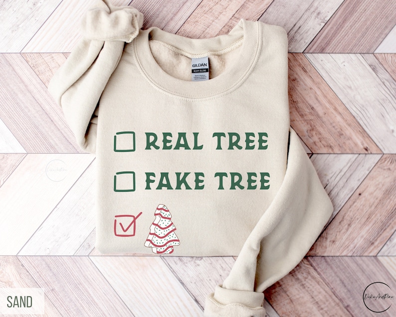 May include: Sand-colored sweatshirt with the text "REAL TREE" and "FAKE TREE" in green, with a checkmark next to a Christmas tree graphic. The sweatshirt is a Gildan brand.