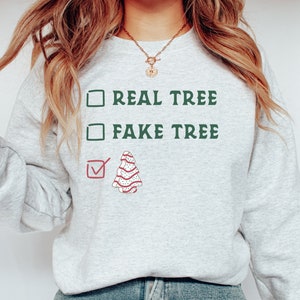 May include: Ash-colored sweatshirt with the text "REAL TREE" and "FAKE TREE" in green, with a checkmark next to a decorated Christmas tree graphic. The sweatshirt is a casual, festive garment.
