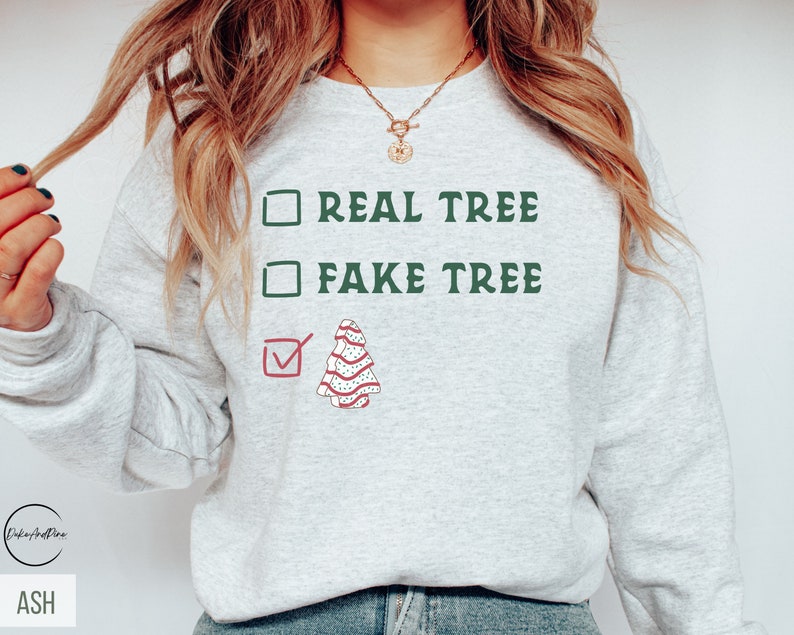 May include: Ash-colored sweatshirt with the text "REAL TREE" and "FAKE TREE" in green, with a checkmark next to a decorated Christmas tree graphic. The sweatshirt is a casual, festive garment.