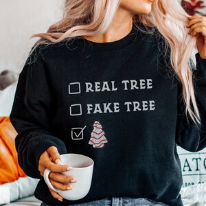 May include: Black sweatshirt with the text "REAL TREE" and "FAKE TREE" in white. A checkmark is next to a graphic of a decorated Christmas tree. The word "BLACK" is at the bottom left.