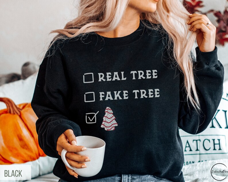 May include: Black sweatshirt with the text "REAL TREE" and "FAKE TREE" in white. A checkmark is next to a graphic of a decorated Christmas tree. The word "BLACK" is at the bottom left.