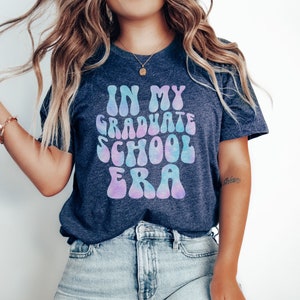 May include: A navy blue t-shirt with a tie-dye effect in shades of pink and blue. The text "IN MY GRADUATE SCHOOL ERA" is printed in a retro font.