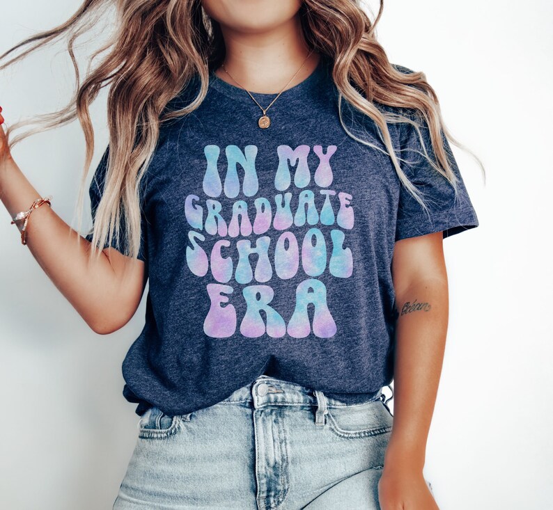 May include: A navy blue t-shirt with a tie-dye effect in shades of pink and blue. The text "IN MY GRADUATE SCHOOL ERA" is printed in a retro font.