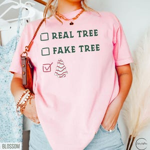 May include: A light pink t-shirt with the text "REAL TREE" and "FAKE TREE" in green, with a checkmark next to a cartoon Christmas tree. The shirt is a casual style.